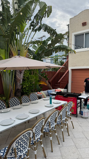 Tropical backyard patio set for outdoor dining — long white table with plates and blue-patterned chairs under an umbrella, string lights overhead, banana palms, and a cook prepping at a portable grill beside a stucco house and garage.