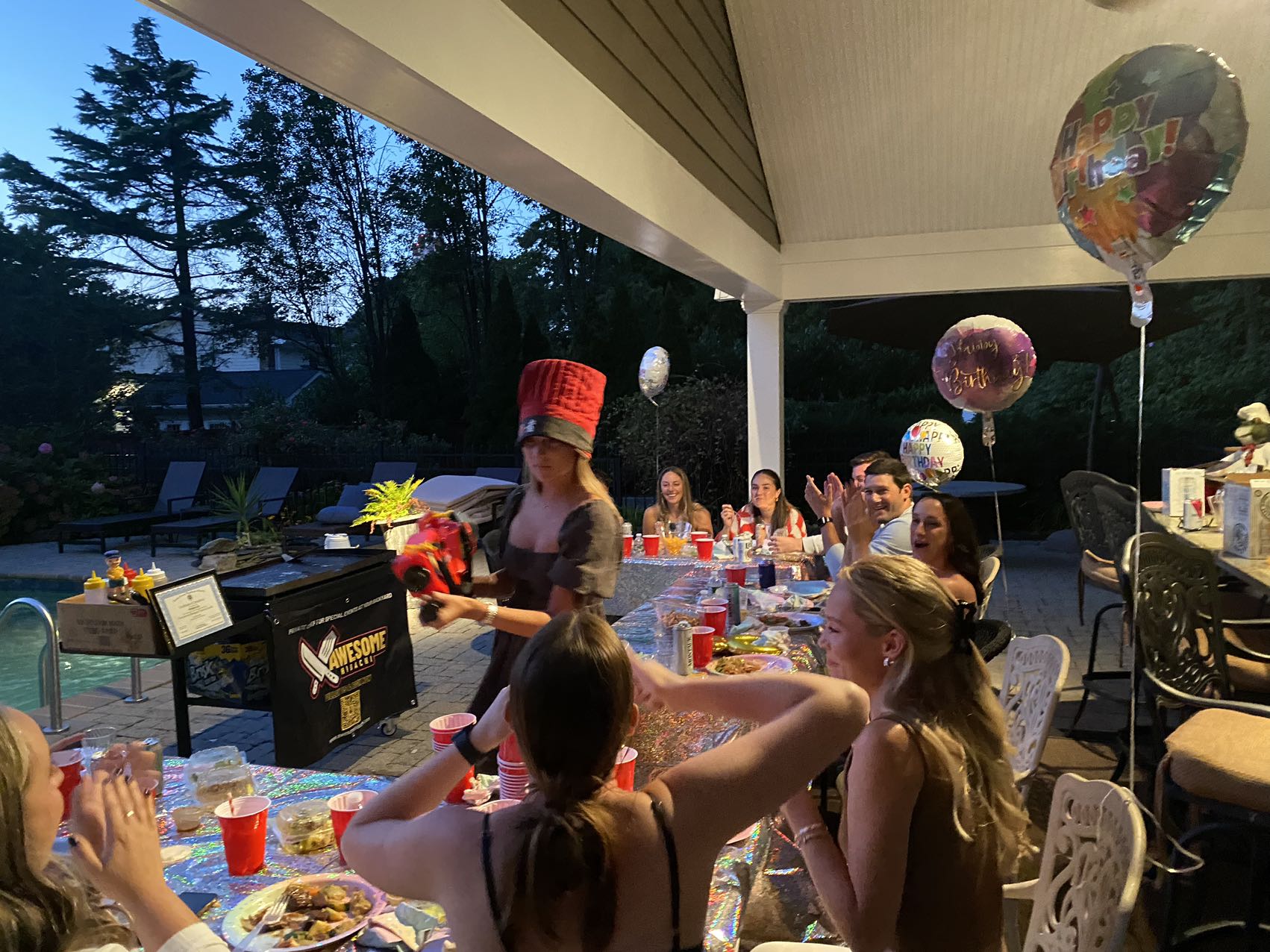 Poolside backyard birthday party on a covered patio at twilight, friends seated at a long sparkly-tablecloth table with red cups and plates, birthday balloons, and a woman in a tall red hat serving food.