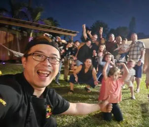 Cheerful backyard group selfie at night — smiling man in foreground wearing glasses and cap with a mixed group of adults and children posing on the lawn under a pergola, casual outdoor family-and-friends gathering.