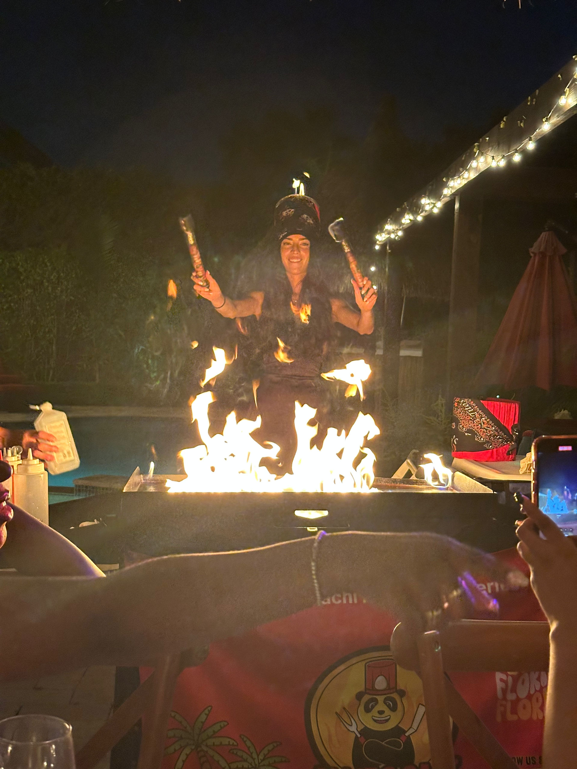 Nighttime outdoor patio fire show with a performer juggling flaming torches over a glowing fire pit, string lights overhead and spectators taking photos.