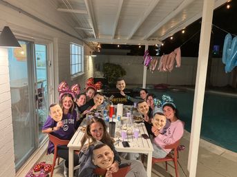 Friends gathered for a backyard poolside patio dinner at night, wearing colorful polka-dot bow headbands and holding oversized printed face cutouts, with string lights and foil balloons overhead.