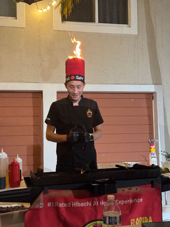 Hibachi chef in black uniform wearing a tall red hat topped by a dancing flame, cooking at an outdoor patio grill with condiments and a water bottle — lively Florida hibachi experience.