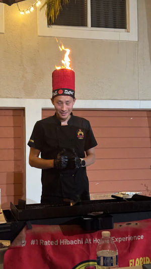 Hibachi chef in black uniform wearing a tall red hat topped by a dancing flame, cooking at an outdoor patio grill with condiments and a water bottle — lively Florida hibachi experience.