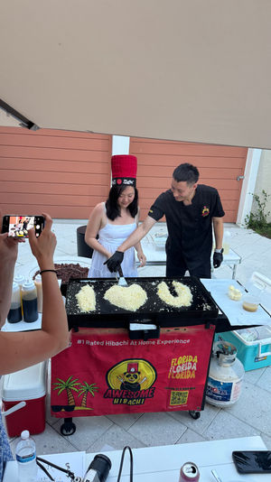 Poolside backyard hibachi in Florida: chef guides a guest at a griddle shaping fried rice into "I ♥ U" while another person snaps a photo.