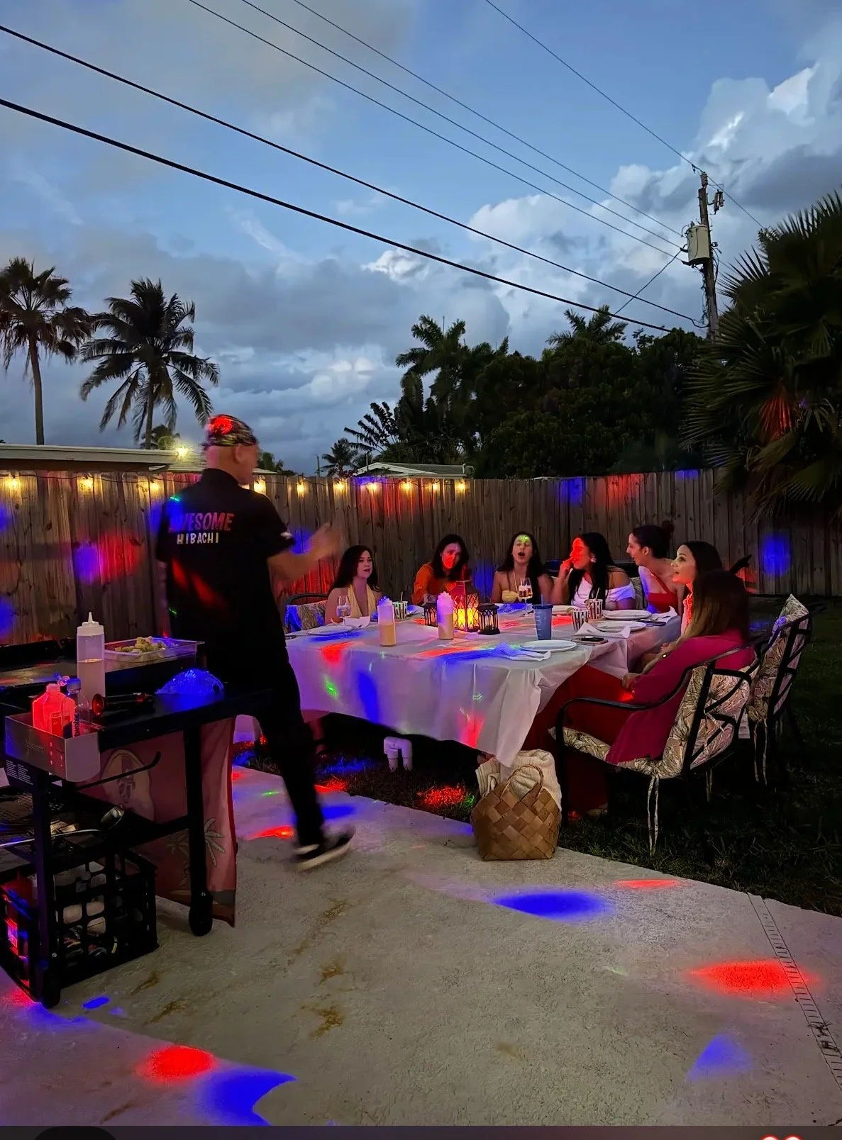Tropical backyard hibachi dinner at dusk with palm trees and cloudy sky, long table of guests under string lights while a chef cooks beside a grill, colorful red and blue party lights reflecting on the table and patio.