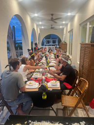 Long covered patio dinner party at dusk on a waterfront porch — long communal table with plates and bowls, arched openings, ceiling fans, and guests in casual clothes and playful headbands