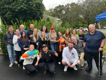 Smiling multigenerational group of family and friends gathered in a suburban front yard/driveway, holding a toddler and a colorful water gun with greenery and an American flag in the background on an overcast day.