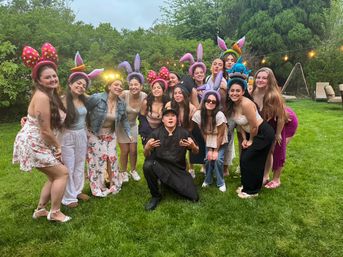 Cheerful group of friends posing at a backyard party on a green lawn at dusk, wearing colorful bunny-eared headbands and summer outfits with string lights and trees in the background.