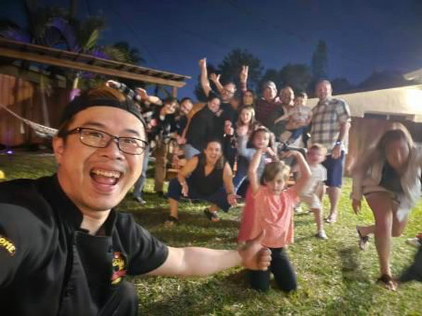Nighttime backyard party selfie: smiling person with glasses in foreground and a lively group of friends and kids on the grass cheering under outdoor lights near a hammock.