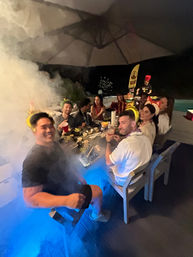 Outdoor poolside patio party at night with a group of friends around a long table, smoky blue lighting, party crowns and leis, plates of food and drinks, and a chef grilling in the background.