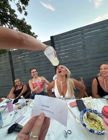 Backyard bachelorette: woman in white leans back as a friend pours a drink from a plastic bottle into her mouth, laughing bridesmaids at a picnic table and a paper slip reading 'The Bride Chicken/Shrimp'.