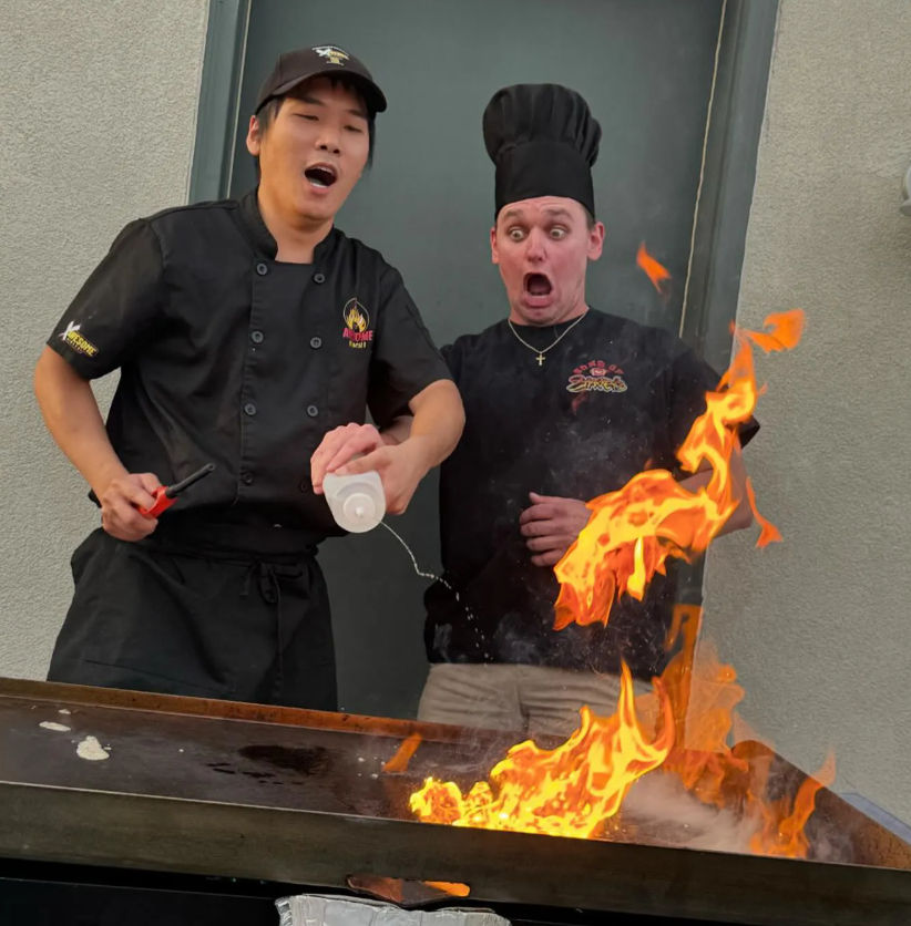 Two chefs at an outdoor teppanyaki griddle create a dramatic hibachi flame show as one pours liquid and both react with surprised expressions.