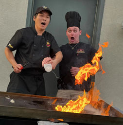Two chefs at an outdoor teppanyaki griddle create a dramatic hibachi flame show as one pours liquid and both react with surprised expressions.