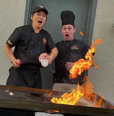 Two chefs at an outdoor teppanyaki griddle create a dramatic hibachi flame show as one pours liquid and both react with surprised expressions.