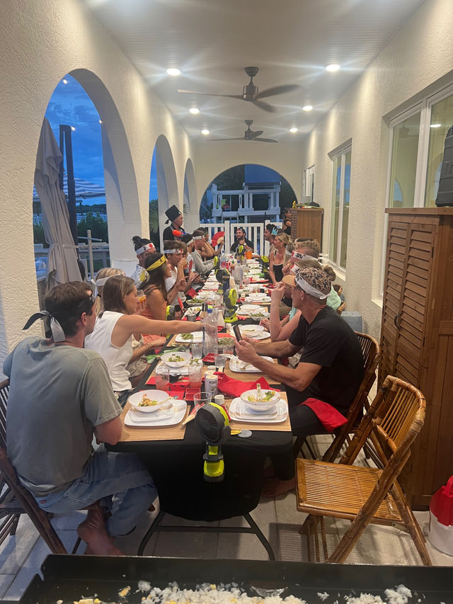 Evening coastal dinner party on a covered arched patio — long communal table with guests wearing bandanas and hats, bamboo chairs, ceiling fans and a waterfront view.