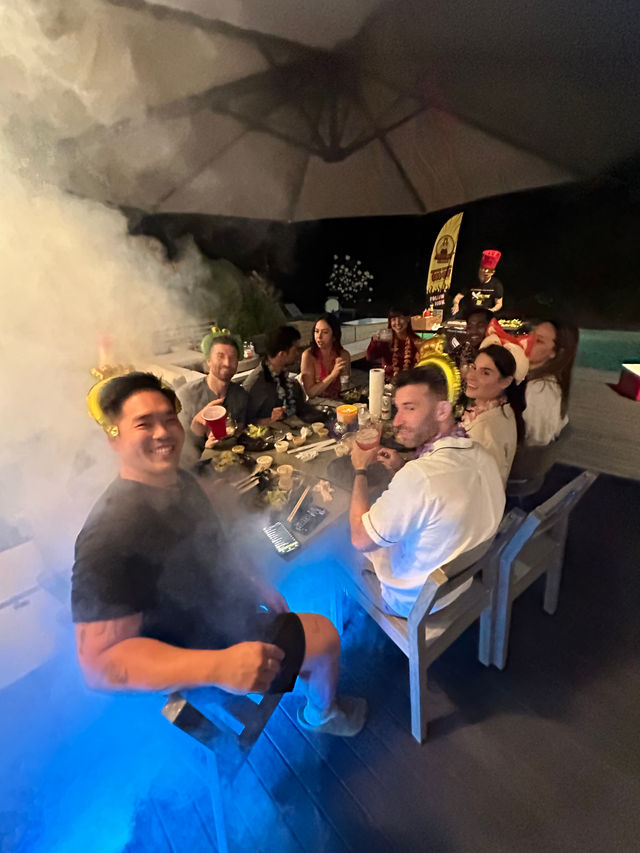 Group of friends enjoying a nighttime poolside patio dinner under a large umbrella, with party hats, drinks, plates of food and smoky blue mood lighting