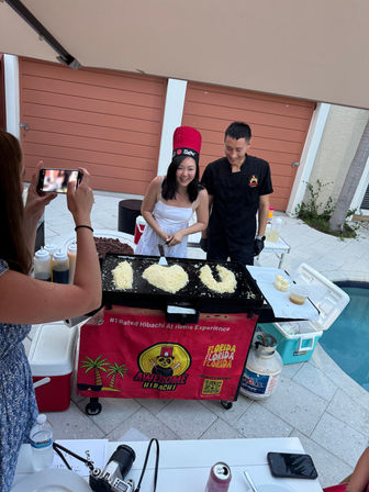 Poolside backyard hibachi: smiling woman in a red chef hat and a chef at a portable griddle, rice formed into "I ♥ U" while a guest photographs them.