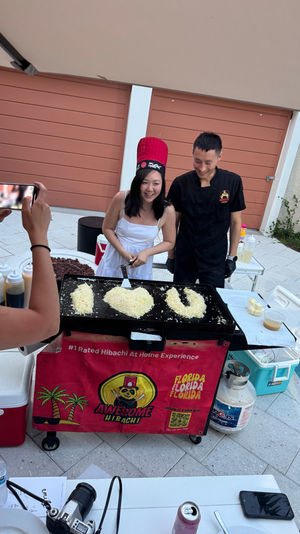 Poolside backyard hibachi: smiling woman in a red chef hat and a chef at a portable griddle, rice formed into "I ♥ U" while a guest photographs them.