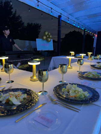 Backyard evening dinner on a covered patio with blue string lights, long white-tablecloth table set with gold goblets, ornate charger plates holding salads, and small gold LED table lamps; host by a grill and a balloon-decorated sign in the background.