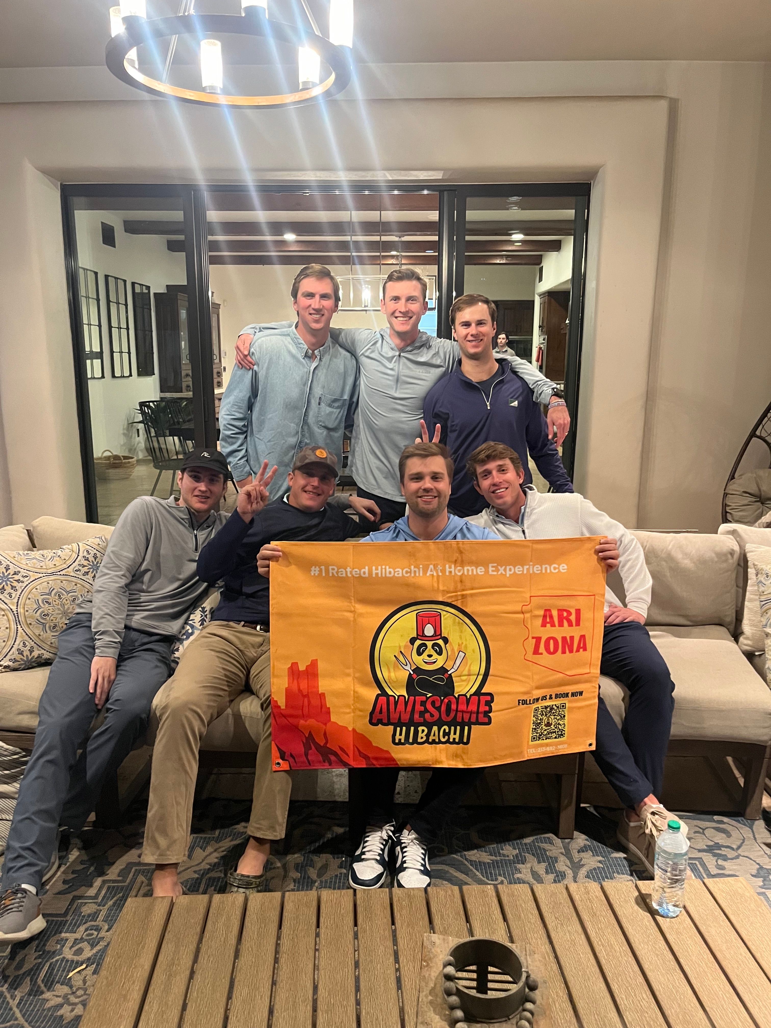 Seven young men smiling in a cozy living room, seated on a couch and holding a bright orange hibachi-style promotional banner with a cartoon chef and the word Arizona, modern chandelier overhead