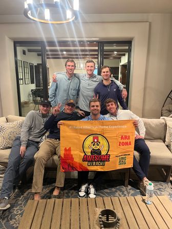 Seven young men smiling in a cozy living room, seated on a couch and holding a bright orange hibachi-style promotional banner with a cartoon chef and the word Arizona, modern chandelier overhead