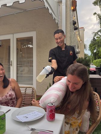 Playful outdoor patio dining scene: smiling server in black uniform playfully squirting a condiment bottle as two laughing diners shield themselves with a striped pillow at a table with plates and canned drinks.