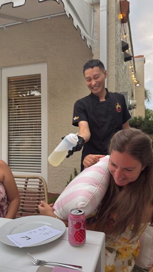Playful outdoor patio dining scene: smiling server in black uniform playfully squirting a condiment bottle as two laughing diners shield themselves with a striped pillow at a table with plates and canned drinks.