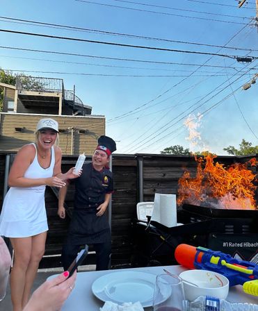 Laughing guest holds a squeeze bottle while a hibachi chef ignites a roaring flame on an outdoor patio grill, plates and drinks in the foreground.