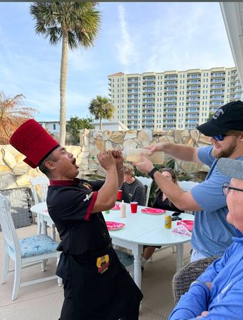 Beachfront patio scene: hibachi chef in a tall red hat playfully opens his mouth as a guest pours sauce from a squeeze bottle, with diners at a white table, palm trees and a high-rise condo in the background.