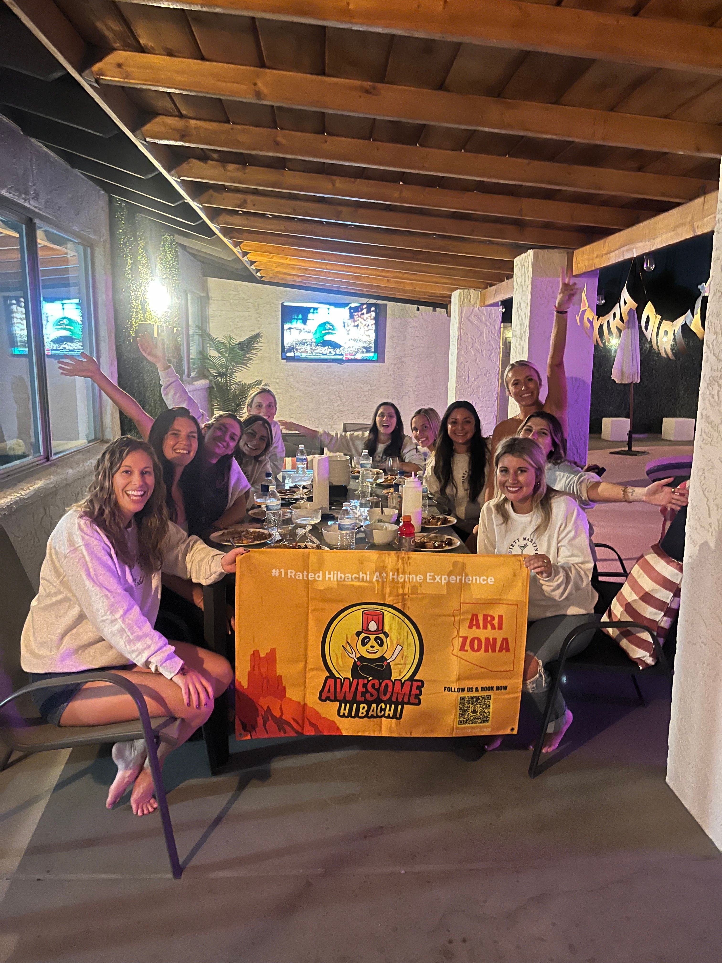 Group of friends enjoying a hibachi-style patio dinner in Arizona, smiling around a long table with plates, drinks and a festive banner under a wooden ceiling.