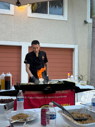 Outdoor hibachi chef cooking teppanyaki on a flat grill with a flaming onion volcano on a backyard patio, sauce bottles and plates of fried rice in the foreground and a palm tree and stucco house behind.