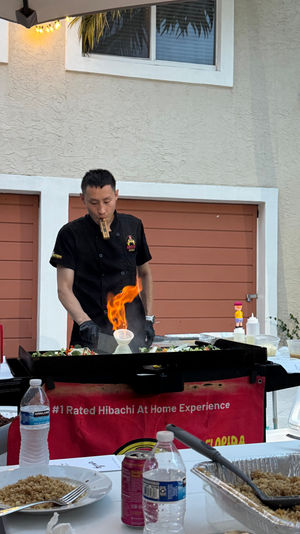Outdoor hibachi chef cooking teppanyaki on a flat grill with a flaming onion volcano on a backyard patio, sauce bottles and plates of fried rice in the foreground and a palm tree and stucco house behind.