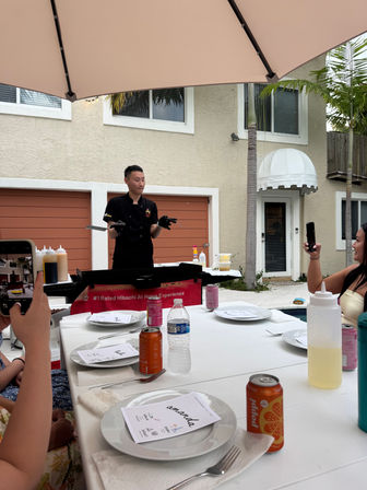 Poolside backyard hibachi chef flipping spatulas at a portable griddle under a patio umbrella while guests photograph the cooking at a white-tablecloth outdoor dining setup with plates, utensils and canned drinks.