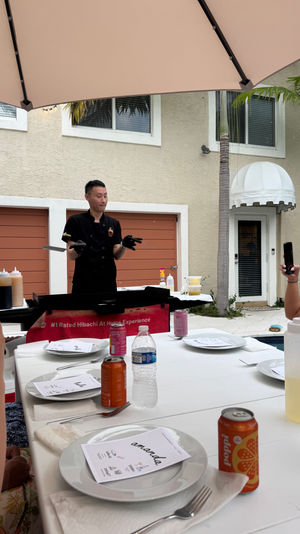 Poolside backyard hibachi chef flipping spatulas at a portable griddle under a patio umbrella while guests photograph the cooking at a white-tablecloth outdoor dining setup with plates, utensils and canned drinks.