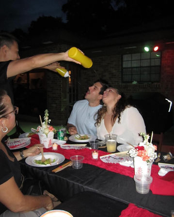 Backyard dinner party with friends — couple leaning back as a person playfully squirts yellow condiment from squeeze bottles into their mouths, table with red runner, takeout boxes and candles.