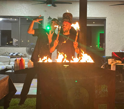 Two performers at a backyard patio party at night, one wearing a tall hat and holding lit torches while both pose behind a flaming grill under string lights and ceiling fans.