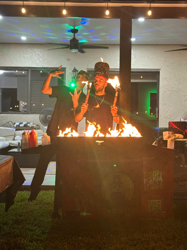 Two performers at a backyard patio party at night, one wearing a tall hat and holding lit torches while both pose behind a flaming grill under string lights and ceiling fans.