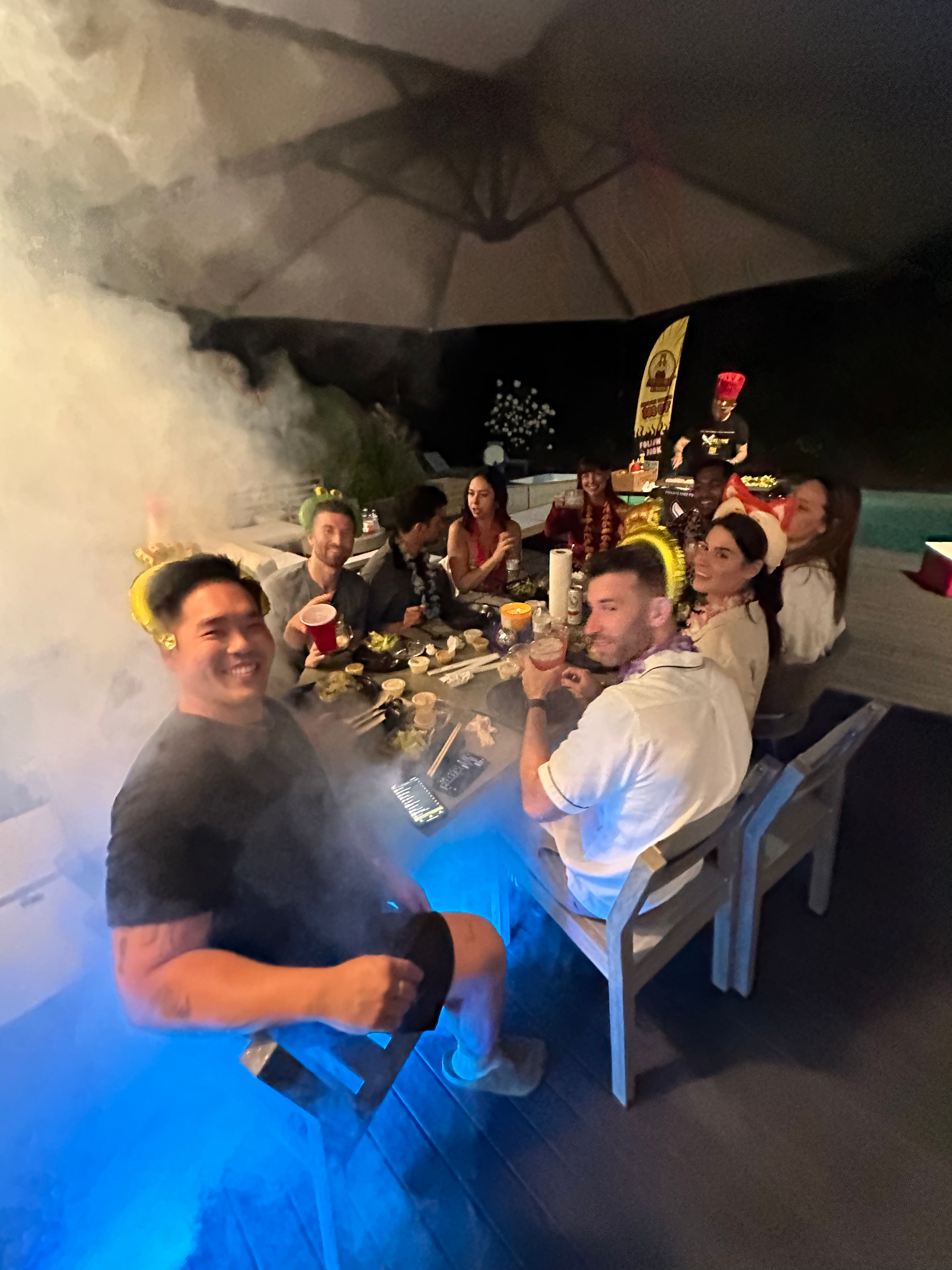 Cheerful group enjoying a backyard poolside dinner party at night on a patio deck — party hats, food and drinks, fog effects and blue mood lighting.