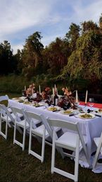 Outdoor garden banquet table with white folding chairs and tablecloth, candlelit floral centerpieces, wine bottle and plated salads on grass beneath leafy trees.