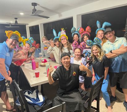 Group of family and friends on a covered outdoor patio at night wearing colorful mouse-ear and bunny-ear headbands, smiling around a table with a birthday cake and drinks, chef giving thumbs-up and festive party vibe.