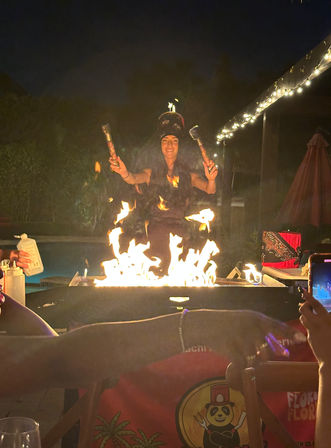 Outdoor nighttime fire show: poolside backyard performer juggling torches above a blazing fire pit, string lights overhead and guests filming on phones.