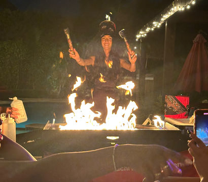 Nighttime backyard fire performance: performer twirling flaming torches over a poolside fire pit on a decorated patio with string lights and spectators photographing the show.
