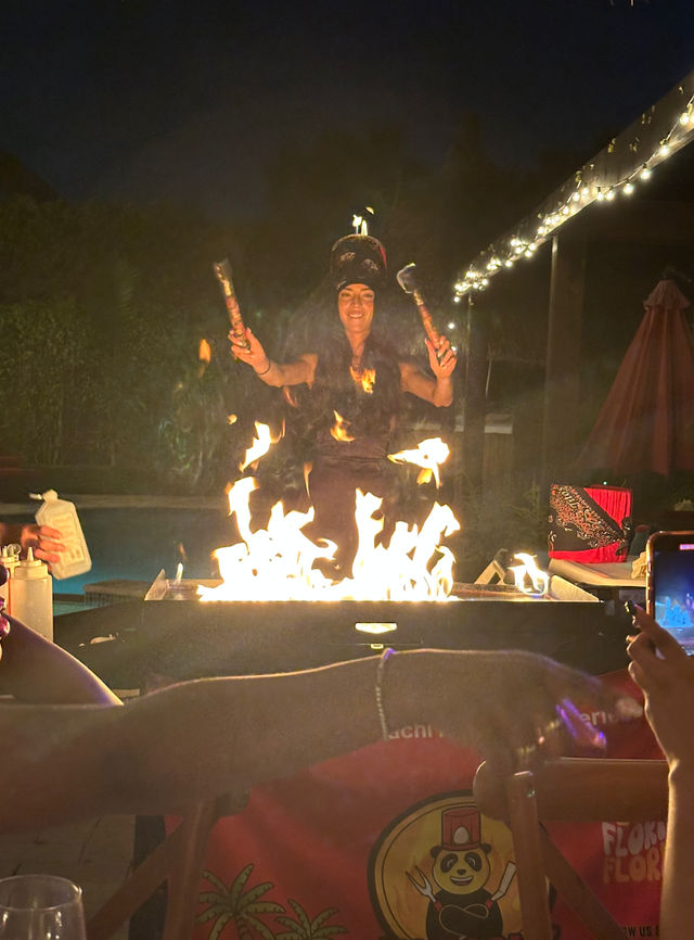 Nighttime backyard fire performance: performer twirling flaming torches over a poolside fire pit on a decorated patio with string lights and spectators photographing the show.