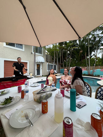 Backyard poolside dinner with three women watching a hibachi chef cook under a large umbrella; white table scattered with plates, soda cans, bottles and condiments, tropical palms and a two-story house in the background.