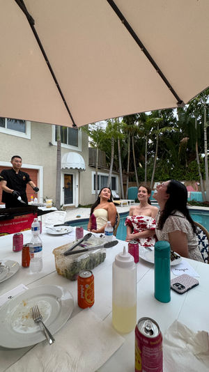 Backyard poolside dinner with three women watching a hibachi chef cook under a large umbrella; white table scattered with plates, soda cans, bottles and condiments, tropical palms and a two-story house in the background.