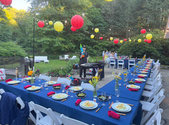Cheerful outdoor backyard garden party on a stone patio with a long blue-tablecloth banquet, white folding chairs, plated salads and red napkins, vases of yellow flowers, and string lights with red and yellow paper lanterns overhead, server by a grill in the background.