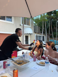 Server playfully squirting whipped cream into a woman’s open mouth at a lively poolside backyard party, friends laughing around a table with trays of rice, drinks, umbrella overhead and tropical palms