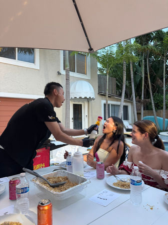 Server playfully squirting whipped cream into a woman’s open mouth at a lively poolside backyard party, friends laughing around a table with trays of rice, drinks, umbrella overhead and tropical palms