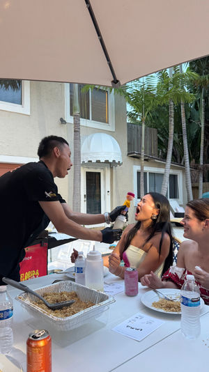Server playfully squirting whipped cream into a woman’s open mouth at a lively poolside backyard party, friends laughing around a table with trays of rice, drinks, umbrella overhead and tropical palms