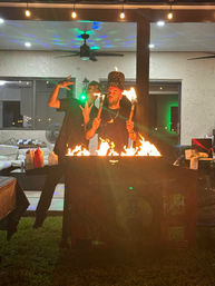 Two performers on a backyard patio at night holding flaming torches over a roaring fire pit, posing playfully under string lights and ceiling fans with outdoor seating in the background.